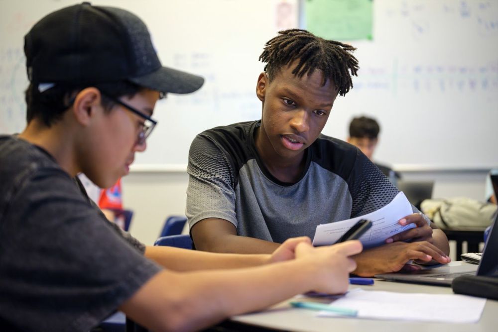 Two students collaborating at a classroom table with papers and notes, discussing and reviewing material